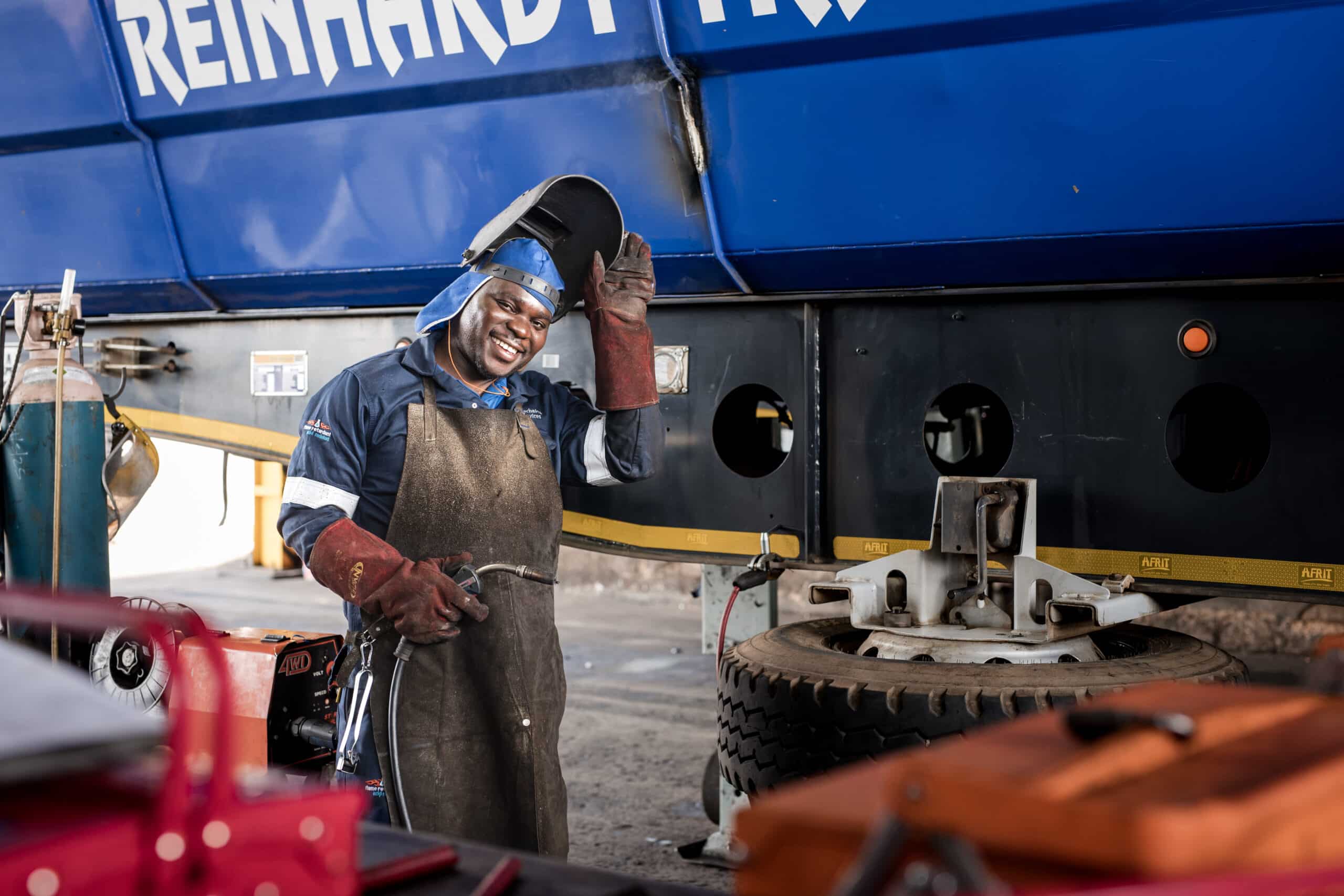 Smiling welder in protective gear standing by a blue trailer
